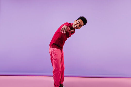 Interested black boy in spring attire pointing finger to camera. Studio portrait of brunette african guy standing on purple background.