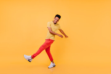 Fototapeta na wymiar Inspired black guy in bright casual attire funny dancing. Laughing african male model in yellow t-shirt and red pants fooling around in studio.