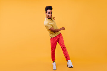 Full-length portrait of glad african guy in red pants fooling around in studio. Indoor photo of curly black man dancing on yellow background.