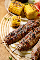Eastern fast food. Grilled spicy beef lyulya kebab on sticks on flat bread with grilled vegetables sweet corn cob, tomato and paprika, tomato sauce on wooden table. Close up
