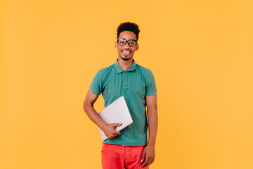 Cheerful international student in green t-shirt smiling on yellow background. Studio portrait of excited male freelancer with laptop.