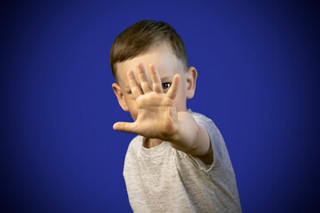 A boy in a gray T-shirt put his hand forward and covered his face with his palm. The child hid behind his hand. Stop gesture. Blue background.