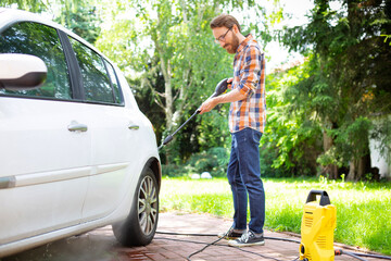 Young man cleaning a car in the garden with a yellow high pressure washer.
