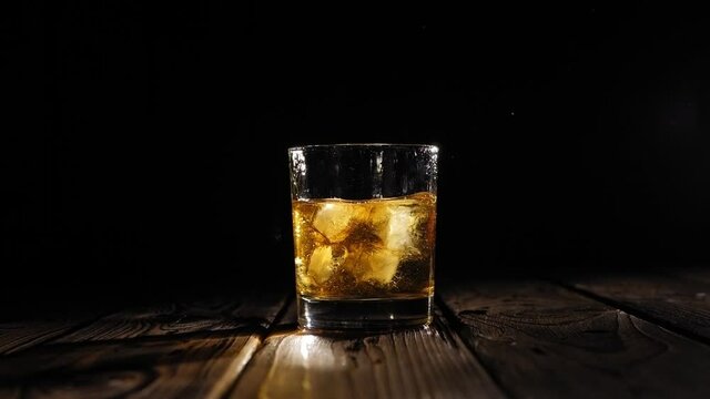Close-up of ice cubes falling into a glass of whiskey on a dark wooden background, a lot of spray flying in different strons. Slow motion. Artificial lighting.