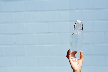 A woman holds a clear glass water bottle in her hand for personal use. On a bright sunny day. Healthy lifestyle and eco friendly reusable and recycle concept. Light blue brick wall background.