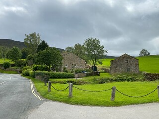 Fototapeta premium House set amongst green fields, with trees, farm building and cloudy skies in, Burnsall, Skipton, UK