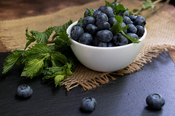 Blueberries with mint leaves in a small white bowl. On a black granite board with a cloth napkin. Wood background.