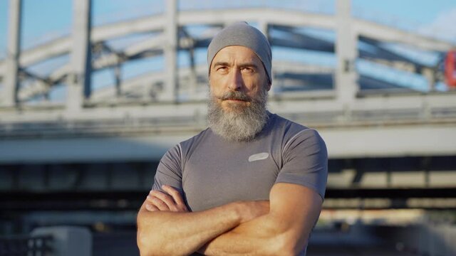 Zoom In Portrait Of Senior Athlete With Grey Beard Finishing Running Workout, Looking At Camera And Crossing Arms Confidently On Bridge Road