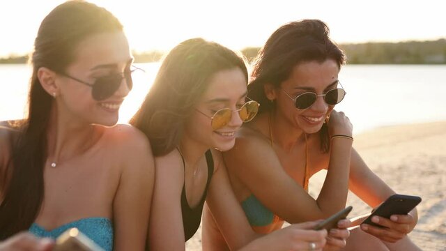 Group of millenial girls using smartphones sitting together on beach towel near sea on summer sunset. Young women addicted by mobile smart phones. Always connected generation communicate via internet.