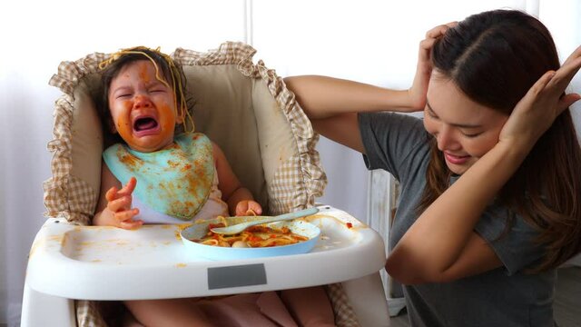 Little Toddler Baby Girl Crying So Loud And Babysitter Get Stress And Use Hands Close Her Ears. Asian Nanny Do Not Know How To Stop Baby Crying, Stubborn Child Get Dirty And Do Not Want To Eat Anymore