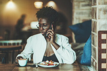 An elegant dazzling young African female in a white trench and fancy eyeglasses is having lunch in a restaurant - she is speaking on the phone and holding a cup of delicious coffee in her hand