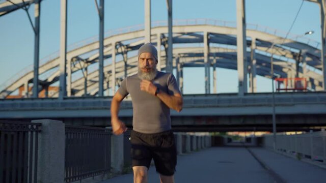 Senior man with grey beard finishing running race, bending forward to catch breath and checking pulse or race time on smartwatch standing on bridge road