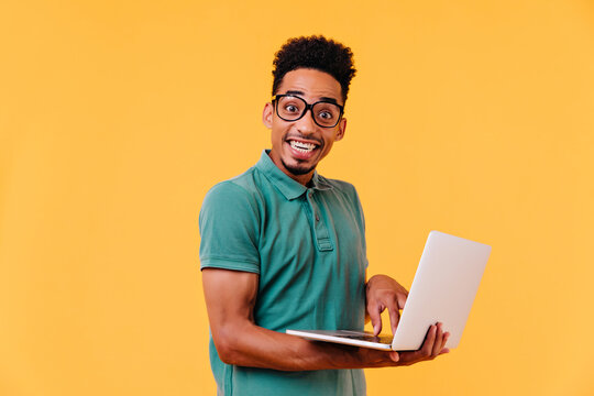 Inspired african student in glasses holding white laptop on yellow background. Studio shot of carefree black male freelancer smiling to camera.