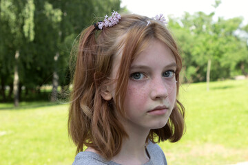 Portrait of a young teenage girl with gray or blue eyes, red hair and freckles, with ponytails on her head. Girl with flowers in her hair in a spring or summer park.