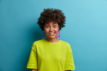 Half length shot of positive ethnic woman reacts on surprising news, bites lips, has shocked glad expression, looks directly at camera, wears green summer t shirt, isolated over blue background