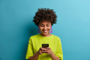 Cheerful pleased woman with curly hair holds mobile phone and texts with friends in social...