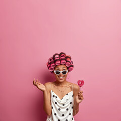 Vertical shot of happy woman concentrated above, raises hand and holds lollipop, wears hair curlers and makes beautiful hairstyle, looks after hair, dressed in polka dot dress and sunglasses