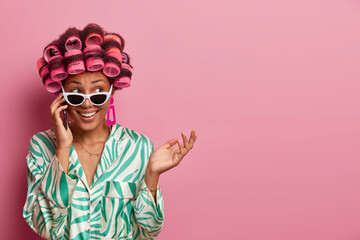 Cheerful hesitant housewife talks on phone, listens rumors and gets pleasant offer wears hair curlers, sunglasses and dressing gown, looks aside happily poses over rosy background blank space for text