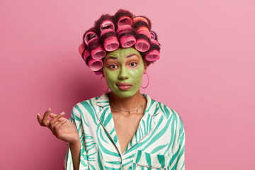 Doubtful perplexed housewife ponders on right decision, shrugs shoulders in bewilderment, dressed in pajama, makes hairstyle with rollers, applies green beauty mask for refreshing skin and complexion