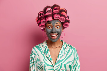 Happy joyful woman visits hairdressing and spa salon, makes perfect hairstyle and applies clay face mask, wears pyjama, has surpised expression, isolated on pink background. Female going on date - obrazy, fototapety, plakaty