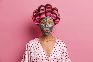 Headshot of beautiful young woman keeps lips rounded, applies beauty mask, wears hair curlers, dressed in casual silk robe, isolated over pink background. Beauty routine and hairstyling concept