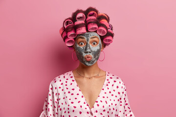 Headshot of beautiful young woman keeps lips rounded, applies beauty mask, wears hair curlers,...
