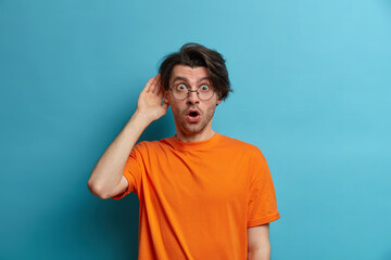 Fototapeta premium People, emotions and lifestyle concept. Surprised amazed man looses speech from wonder, stares with bugged eyes and opened mouth, hears terrible news, wears orange t shirt, transparent glasses