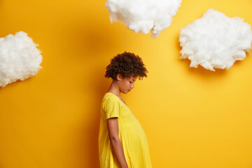 Profile shot of African American woman with pregnant belly, looks down on her tummy, wears yellow...