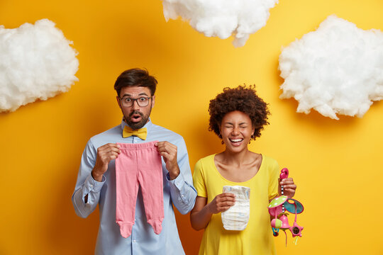 Married couple expect for child. Husband and wife pose with baby stuff, Afro American pregnant woman laughs happily, holds diaper and mobile, shocked future father poses with newborn clothes - Powered by Adobe