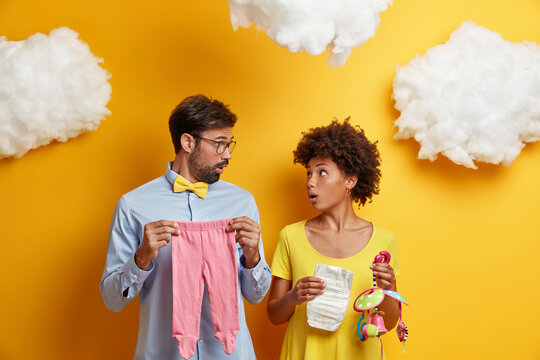 Shocked husband and wife await for little newcomer, buys necessary items for baby, stare at each other, enjoy home atmosphere, stunned to confuse gender of future child. Expectant parents indoor - Powered by Adobe