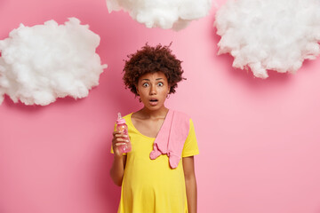 Shocked emotional pregnant woman dressed in yellow dress, has round belly, poses with baby items, realizes two weeks left before delivery, almost nine months of exciting expectations behind.