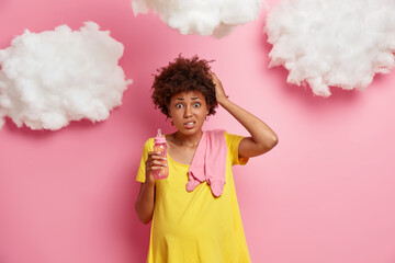 Puzzled pregnant woman has no experience of motherhood, looks worried at camera, holds baby bottle with nipple, doesnt know how to care about newborn, expects new life, has confused expression