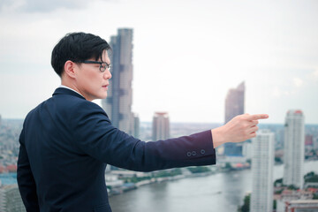 Young Asian businessman in suit pointing to the sky with skyscraper city