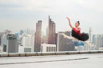 Fototapeta na wymiar Asian ballerina dancer girl jumping in the air, practicing ballet dancing on rooftop with skyscraper city view, adorable child dancing in ballet