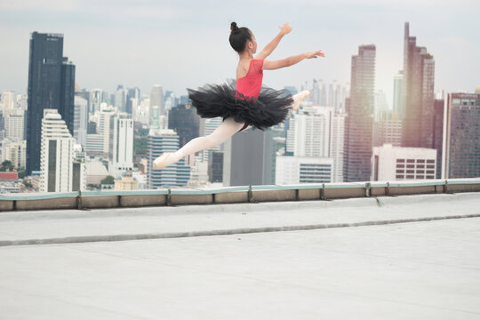 Asian ballerina dancer girl jumping in the air, practicing ballet dancing on rooftop with skyscraper city view, adorable child dancing in ballet
