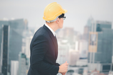 Happy Asian young businessman engineer in suit wearing safety helmet, singing, and dancing on the rooftop with skyscraper city view