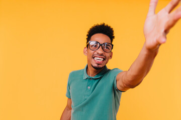 Confident african guy looking up and waving hand. Indoor portrait of curly male model in big glasses.