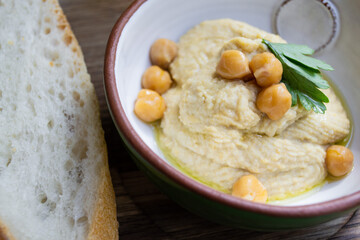 Hummus decorated with parsley and chickpea peas in a ceramic bowl with a piece of bread on a wooden background