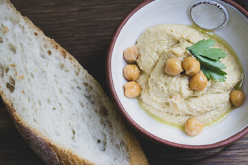 Hummus decorated with parsley and chickpea peas in a ceramic bowl with a piece of bread on a wooden background