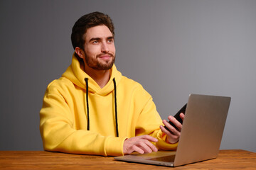 A young guy with different emotions in a yellow sweatshirt works using a laptop and smartphone. 