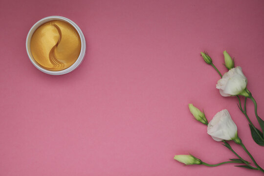 top view of golden yellow cosmetic spa eye patches in white jar and white flowers with green leaves on pink background, flat lay