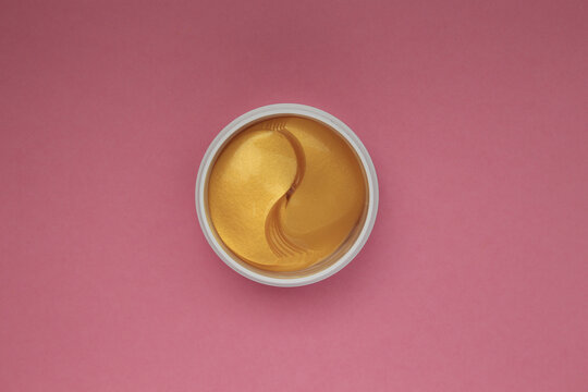 top view of golden yellow cosmetic spa eye patches in white jar on pink background, flat lay