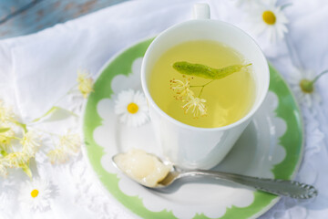 Linden flowers tea in a white cup on a white with green ornament plate with spoon with honey on a blue wooden background and white napkin outdoors, yellow drink of linden flower on  vintage background