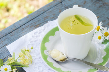 Linden flowers tea in a white cup on a white with green ornament plate with spoon with honey on a blue wooden background and white napkin outdoors, yellow drink of linden flower on  vintage background