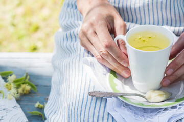 Woman keeps white cup with fresh linden flowers tea and plate with spoon and honey on a stripped apron background outdoors, person rests on a blue colored bench in the garden