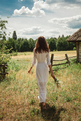 A girl in a white dress holds a bouquet of daisies in her hands. Girl outdoors in the village, against the background of the field.
