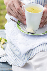 Woman keeps white cup with fresh linden flowers tea and plate with spoon and honey on a stripped apron background outdoors, person rests on a blue colored bench in the garden