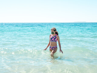 Young girl on the beach wearing a protective mask to avoid coronavirus disease, safe holidays concept