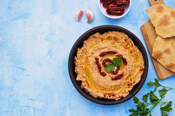 Hummus with sun-dried tomatoes in a brown clay bowl on a light blue concrete background. Served with tortillas, drizzled with olive oil when applying. Healthly food. Recipes chickpeas.