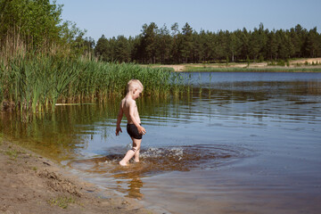 blond boy in black swimming trunks neatly enters the lake with yellow sand, in the background coniferous forest.
lmage with selective focus